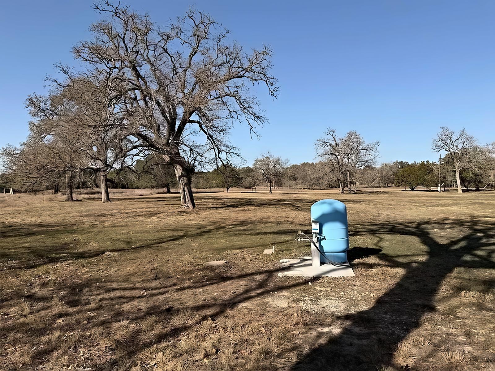 Blue well pressure tank on a concrete pad in the meadow, with oaks behind it